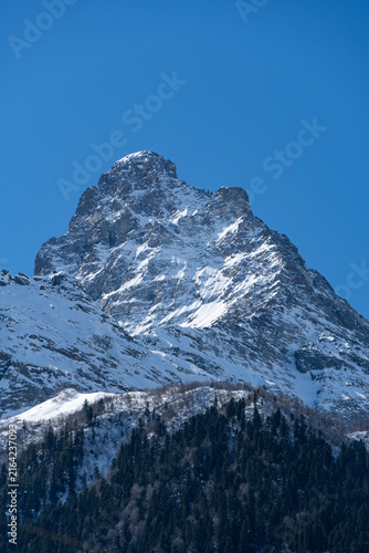 Majestic snow-capped mountain peak stands tall against clear blue sky, with rugged terrain and patches of snow covering its slopes. Karachay-Cherkessia. Dombay 2025. Nature concept for design.