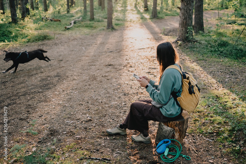 Woman using smartphone while resting during a dog walk on a forest trail with backpack and leashes nearby