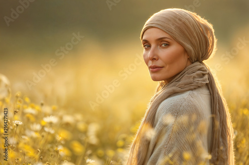 A woman wearing a draped beige headscarf and knit cardigan stands in a meadow of white daisies, glancing back at the camera as warm golden light backlights the field.