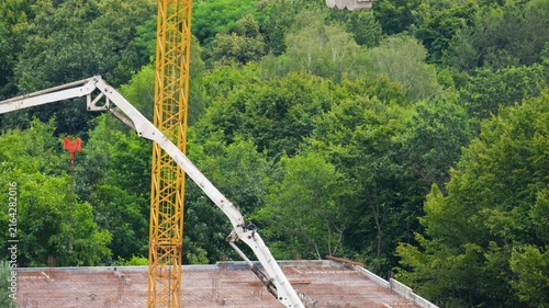 A high-angle view of a tower crane next to an unfinished modern building surrounded by green trees, as a concrete pump arm pours cement mixture and workers prepare reinforced metal mesh and formwork.