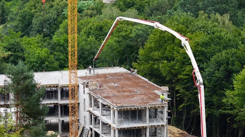A high-angle view of a yellow tower crane and concrete pump truck working near a forest, as safety-vested workers control a hose to pour mixture into formwork on a multi-story building.