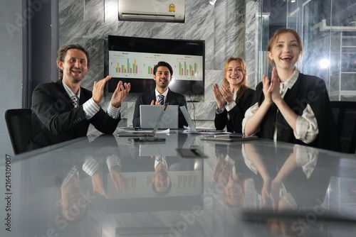 Obraz na płótnie Dark haired executive cups his hands mid gesture while colleagues laugh and clap, spontaneous joy rippling through the nighttime glass walled office in a bright modern coworking office
