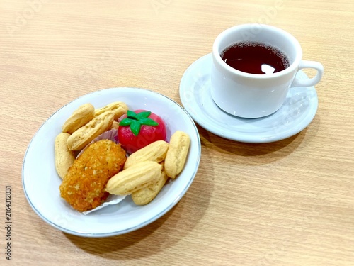 A white plate holds an assortment of traditional Indonesian snacks including cookies, savory fried croquettes, and a sweet red, tomato-shaped treat accompanied by a cup of strong tea with a saucer.