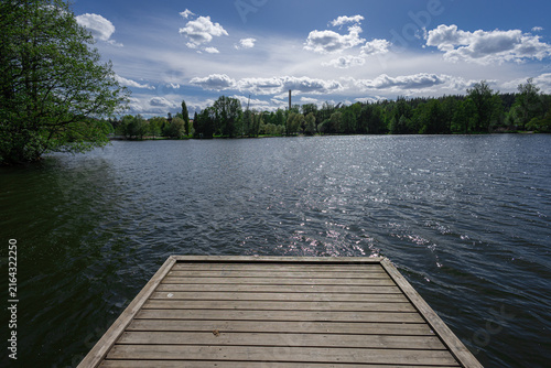 A weathered wooden dock extends over a sparkling lake in Lahti, framed by lush green trees and a bright blue sky dotted with fluffy clouds on a sunny summer day.