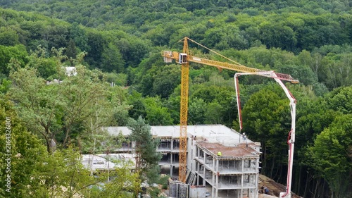 A high-angle view of a yellow tower crane and concrete pump truck working near a forest, as safety-vested workers control a hose to pour mixture into formwork on a multi-story building.