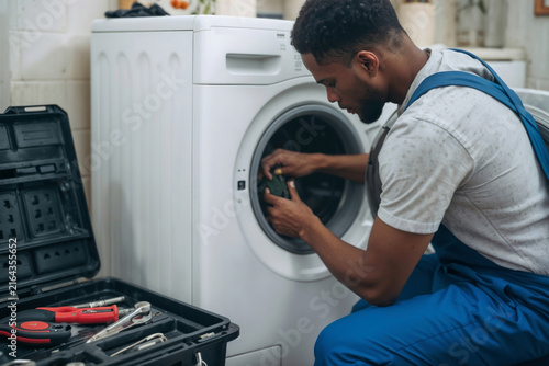 Skilled technician fixes washing machine with care. Tools rest open beside him, ready for use. Focused, he works through machine's door. Routine repair scene, calm and professional