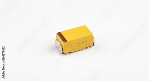 Macro photograph of a single bare SMD tantalum capacitor. Yellow epoxy casing with polarity stripe and metallic mounting tabs. Electronic hardware isolated on a pure white background cutout.
