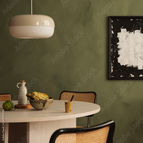 Modern minimalist dining room interior with round wooden table, rattan chairs, pendant lamp, and abstract art piece against a dark green wall