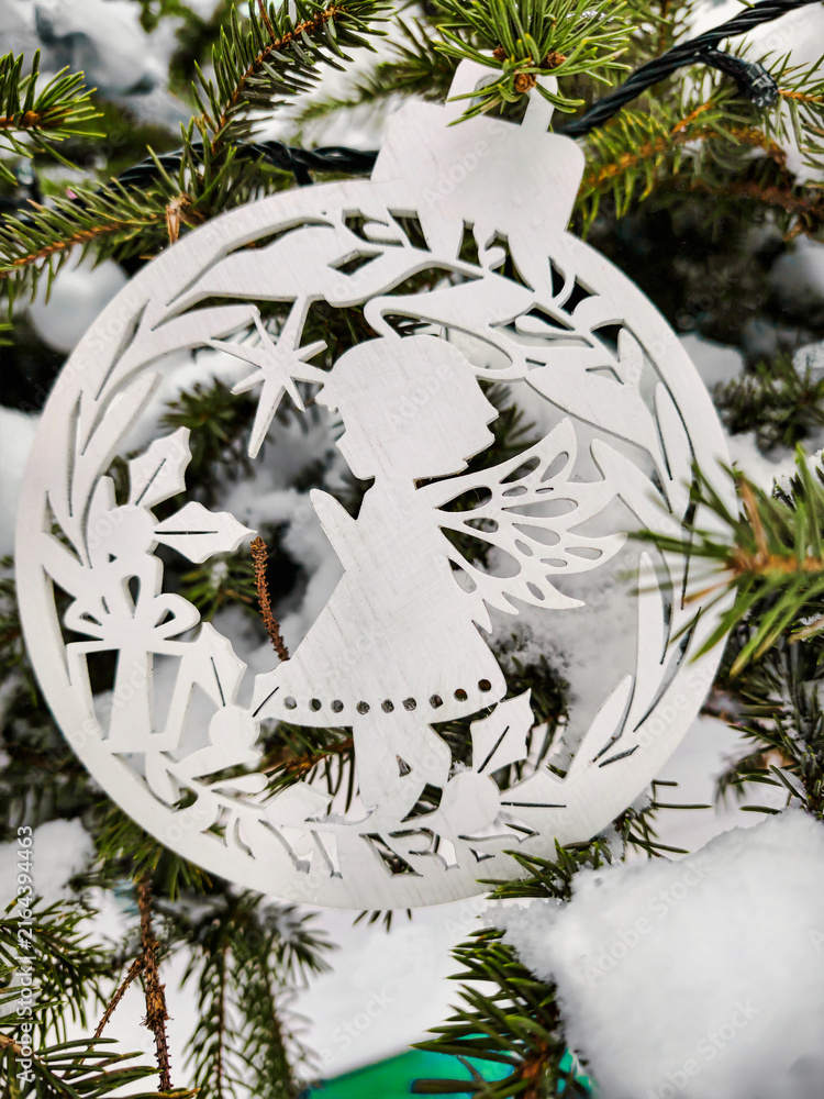 Fototapeta premium A white ornament shaped like an angel hangs on a Christmas tree. Snow covers the ground and tree, creating a festive atmosphere for the holiday season