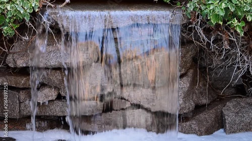 A close-up shot of a small, decorative outdoor waterfall cascading over a wall of stacked grey stones into a bubbling pool of foam, framed by green ivy.