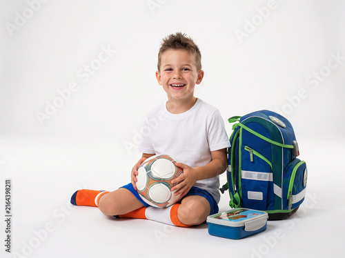 Smiling little boy in blank white t shirt holding soccer ball sitting near school backpack and lunchbox isolated on white background with copy space for design or text