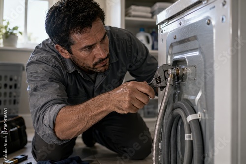 Male technician in gray shirt and pants repairs washing machine plumbing connections with adjustable wrench in a laundry room, surrounded by cleaning supplies and laundry baskets