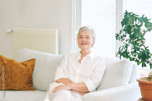 55-60 year old woman sitting comfortably on sofa at home, smiling mature female in white clothes relaxing in bright living room, confident and peaceful lifestyle