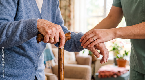 Caregiver supporting elderly person holding walking cane indoors with gentle handhold