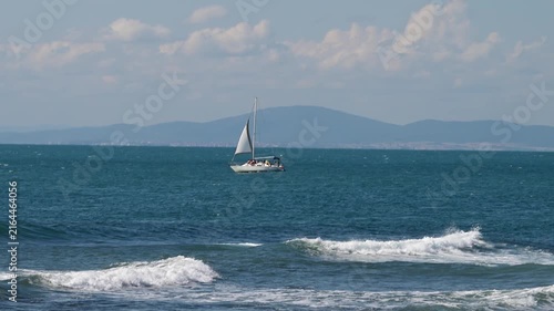 A white sailboat cruises across a vibrant blue sea with rolling waves and whitecaps, set against a backdrop of distant mountains and a partly cloudy sky.