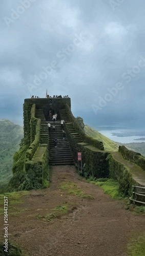 Photography Trekkers climbing the steep stone staircase of an ancient hill fort bastion overlooking a lush valley and lake in the Western Ghats, Maharashtra