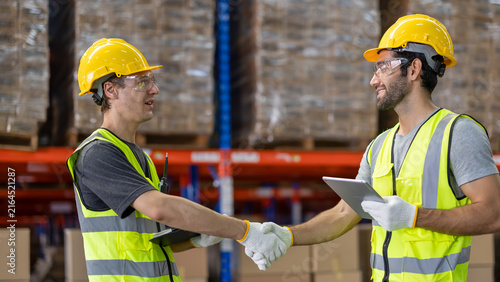 Warehouse workers shaking hands in a large fulfillment center.Logistics team members handshake greeting in a storage facility.Two diverse industrial engineers shaking hands in a warehouse.