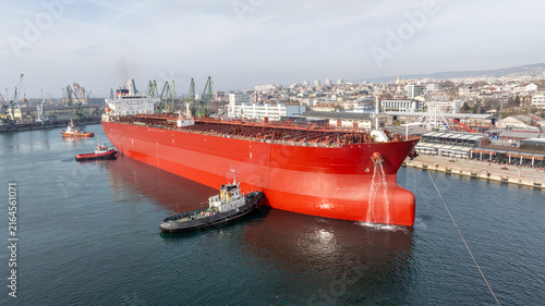Aerial View of a Large Red Oil Tanker Assisted by Several Tugboats in a Busy Industrial Port Harbor with an Urban City Background