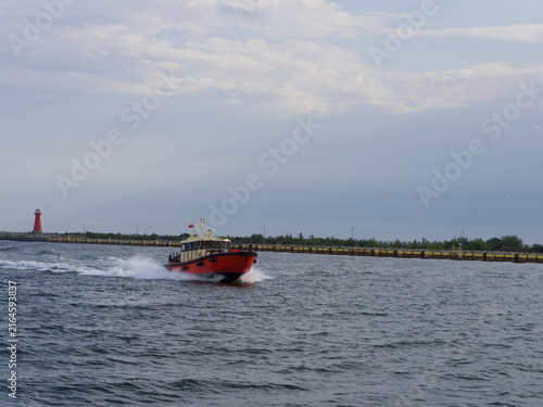 Photography Boat moves swiftly through water near a harbor with a lighthouse and cloudy sky