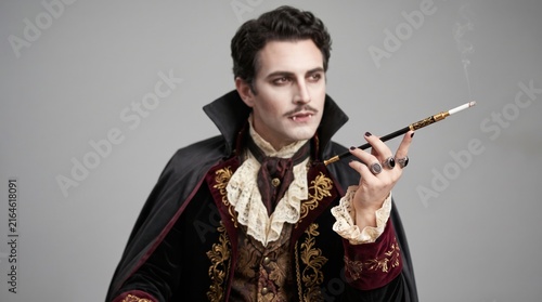 A man dressed as a Victorian-era vampire smokes a cigarette with a long holder, wearing an ornate coat and cape against a plain grey background.