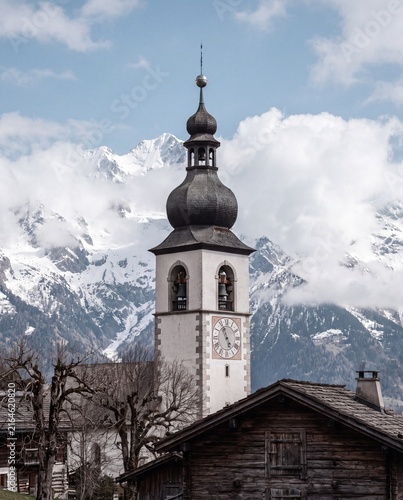 Picturesque alpine village church tower with a clock against a dramatic backdrop of majestic snow-capped mountains and clouds