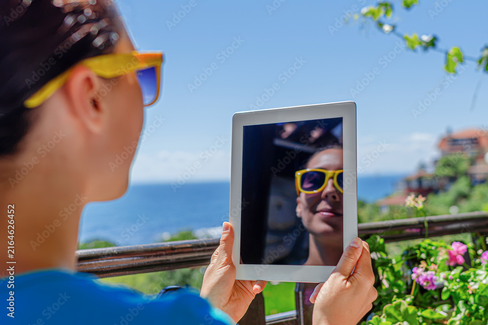 Fototapeta premium A young woman wearing sunglasses holds up a digital tablet that reflects her smiling face on a sunny balcony overlooking the sea.