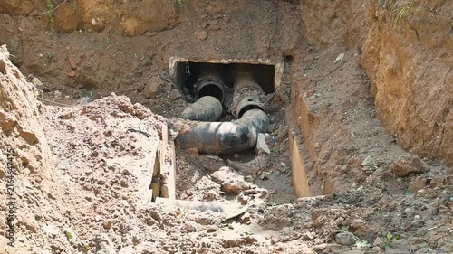 excavation work to install or repair underground pipes at a construction site. The location is a dirt area with deep trenches revealing large pipes and heavy machinery
