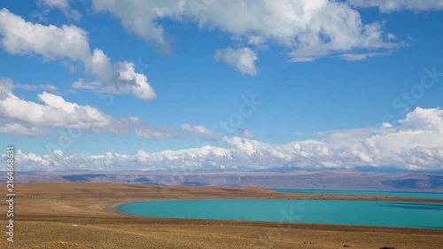 Turquoise glacial lake surrounded by golden steppe hills under a dramatic blue sky with fluffy white clouds in remote highland landscape