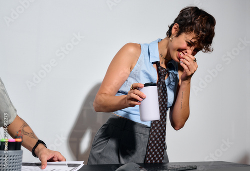 Woman laugh at desk in business office hours. A woman at her desk is visibly happy. She holds a mug while laughing,. Her posture and expression convey hapiness.  Direct flash. Business office.
