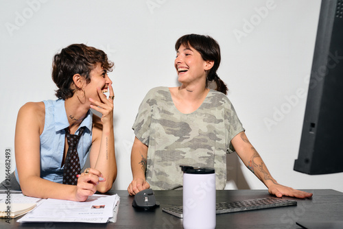 Two women share a joyful laugh at their desk. They’re engaged in a lighthearted moment, hands near their faces as they smile.