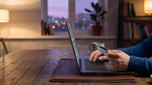 Man holding credit card while shopping and making payments online on laptop at home in evening