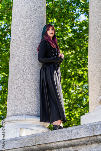 Young woman with colorful hair wearing a black gothic dress and boots, posing between monumental stone columns surrounded by green trees outdoors.