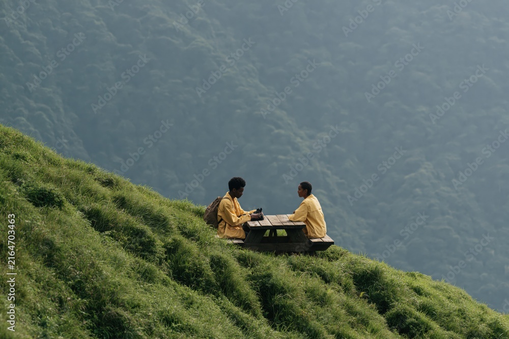 Fototapeta premium Two people sitting at picnic table on steep grassy hillside in matching yellow jackets for travel background and outdoor adventure banner