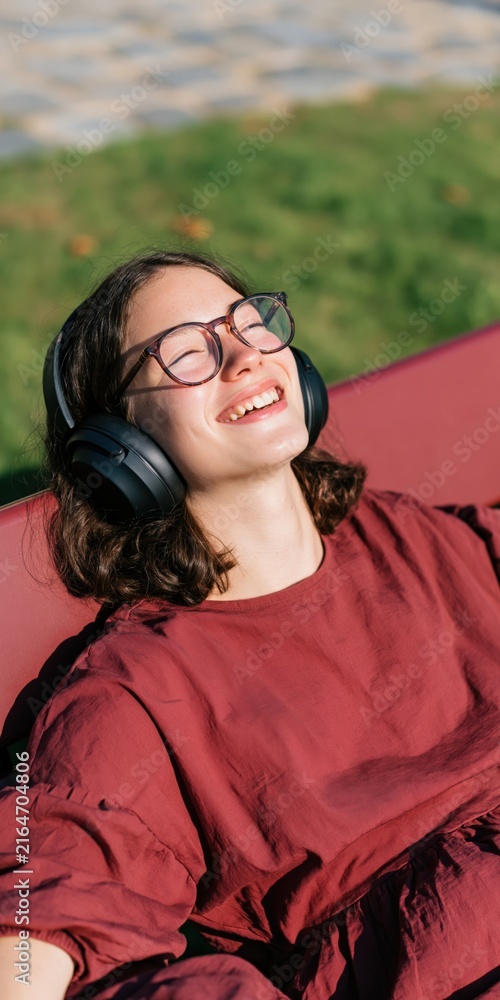 Naklejka premium Smiling young woman wearing headphones outdoors relaxing on a bench in sunlight for banner background and portrait design