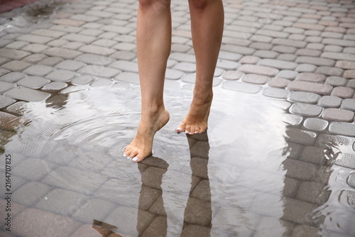 Obraz na plátně Close-up of a woman standing barefoot in a puddle on a wet cobblestone street after rain
