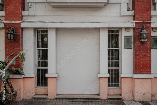 Canvas Print Ground level exterior of a building featuring a blank white central wall space, flanked by narrow barred windows, red brick pillars, vintage-style wall lanterns, and a potted plant on a paved walkway