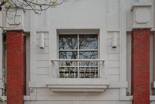 Photography Detailed view of a white building exterior featuring a glass window with tree reflections, a decorative white metal balcony railing, symmetrical red brick pillars, and cylindrical wall lamps