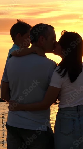 Family enjoying sunset at beach. Parents share loving kiss while child observes. Warm atmosphere creates joyful memories. Perfect vertical scene for family moments.