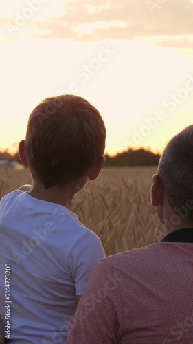 Father and son stand in wheat field watching sunset together. Little boy points toward distant horizon as both dream about future. Golden evening light reflects trust, guidance and family values.