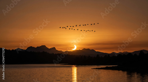 A scenic view of a solar eclipse over mountain range with migrating birds flying above a tranquil lake during sunset