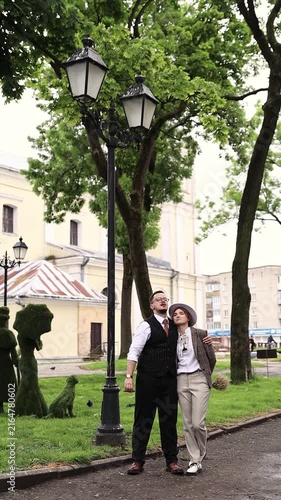 An eccentric couple standing on a park path near a vintage lamppost, with the groom smoking a cigar. Groom smoking a cigar, unique vintage bride fashion, nonconformist wedding concept, and elegance