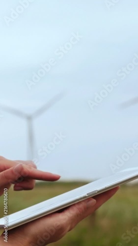 Person using tablet outdoors near wind turbines. Modern technology supports renewable energy awareness. Green energy concept in vertical format. Nature and innovation unite.
