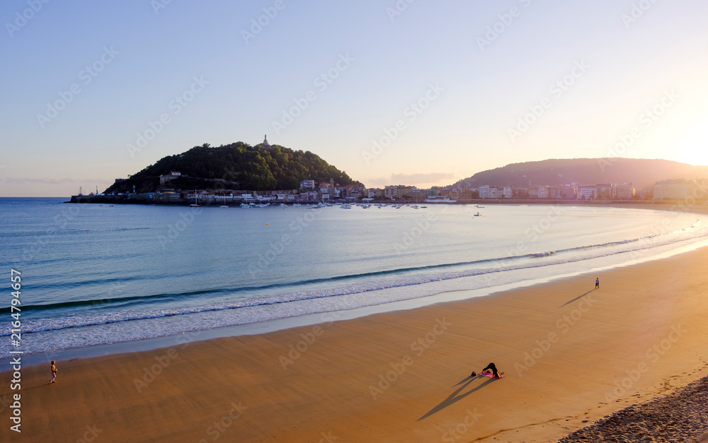 Fototapeta premium Couple walking at sunrise in La Concha Bay, San Sebastian Donostia, Basque Country, Spain. Scenic view of Urgull mountain and city skyline.