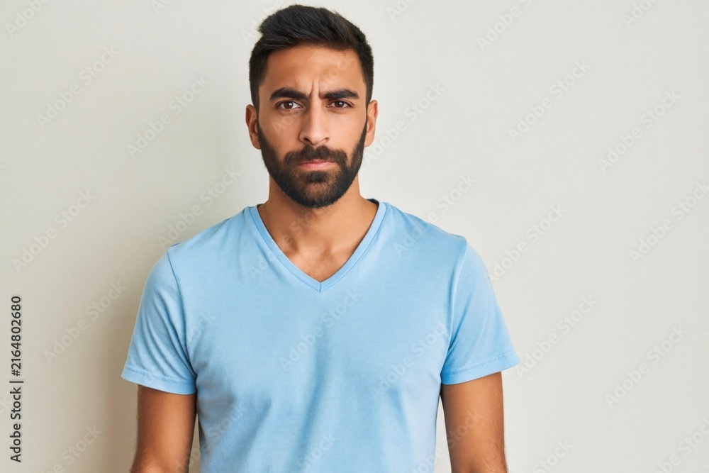 Obraz premium Young man with beard expresses doubt and distrust. His raised eyebrow and serious look show uncertainty. He stands indoors against a light gray background.