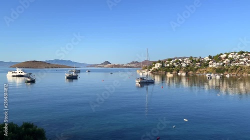 Scenic view of the calm, blue bay in Bitez, Turkey, with luxury boats anchored on the still water near a hillside resort town under a clear morning sky.