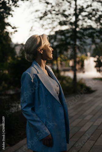 Elegant blonde woman with light skin and refined makeup wearing a blue suit, posing in summer sunlight with artistic shadows and dramatic light, creating a sophisticated fashion and luxury portrait