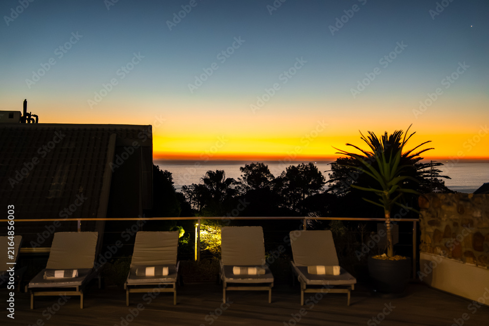 Fototapeta premium Sun loungers with folded towels line a hotel terrace facing the sea horizon during a vibrant evening sunset in Cape Town, South Africa.