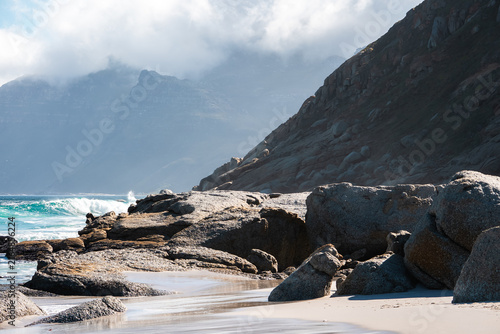 Large boulders and sandy shore border the turquoise sea with mountains rising into cloudy skies along the coast in Cape Town, South Africa.