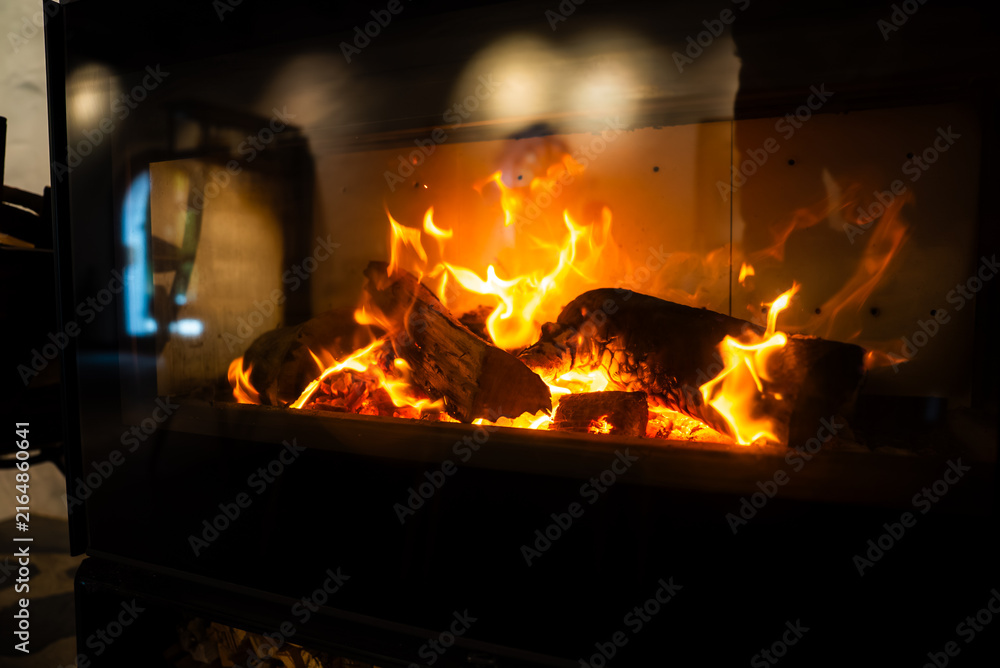 Fototapeta premium Bright orange flames engulf burning wood logs within a glass enclosed indoor fireplace, generating comforting warmth and atmospheric interior light.