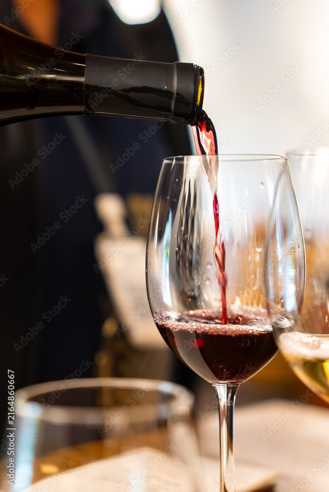 Naklejka premium Dark red wine pours smoothly from an open dark bottle neck into a stemmed clear glass during an indoor drink tasting session.